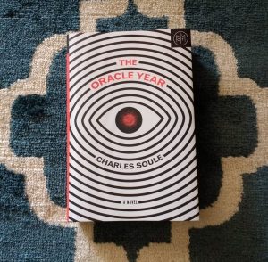 Photo of the cover of The Oracle Year by Charles Soule. Cover features a black and white eye with a red iris.